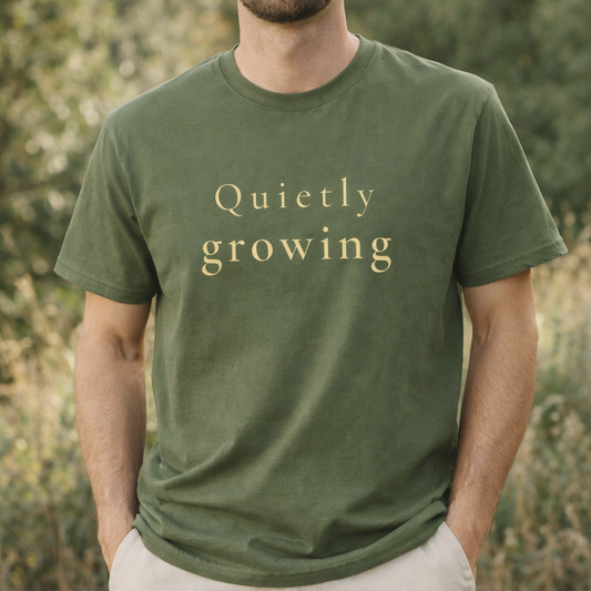 male model wearing sage green Comfort Colors T-shirt with “Quietly growing” text, relaxed outdoor lifestyle image with natural light