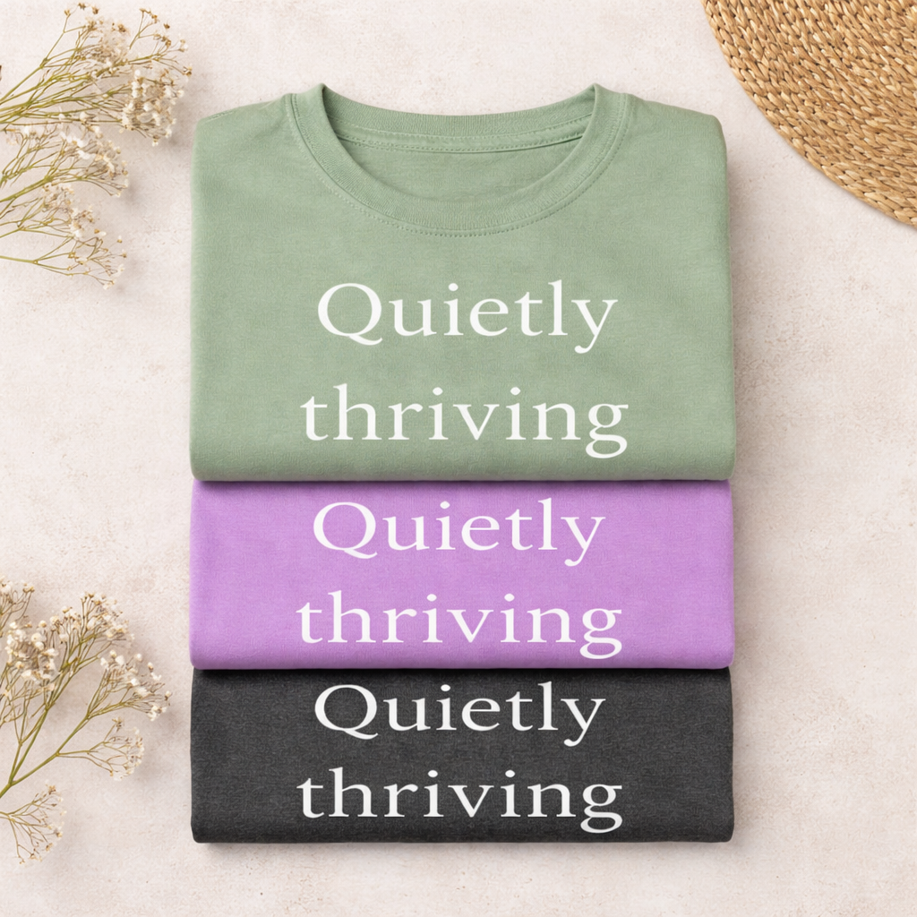 folded stack of Bella Canvas T-shirts in sage green, heather team purple, and dark grey heather with “Quietly thriving” text, styled on a neutral surface with soft textures
