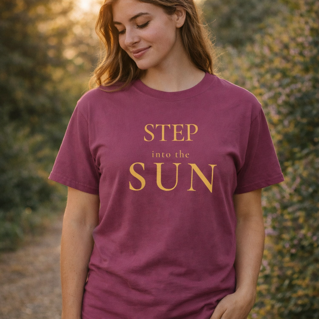 Woman wearing berry “Step Into the Sun” T-shirt outdoors in soft sunlight, relaxed expression looking down, warm natural setting with greenery