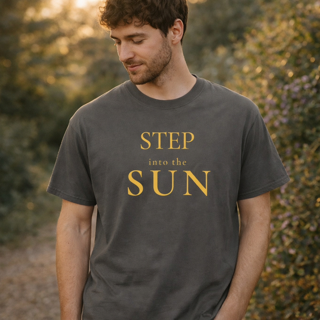 Man wearing charcoal “Step Into the Sun” T-shirt standing outdoors in warm golden light, relaxed posture with hands in pockets, soft greenery background