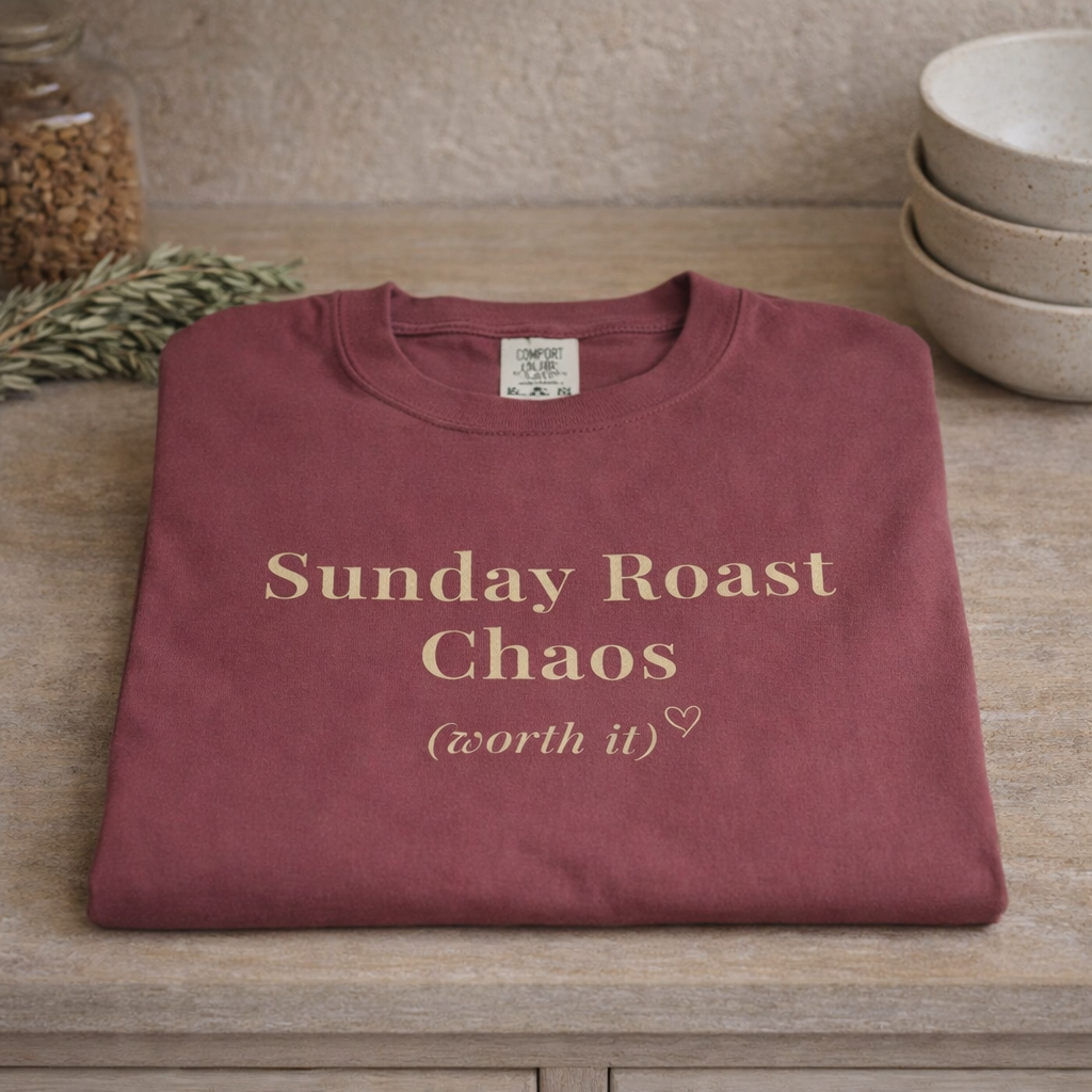 Folded burgundy Sunday Roast Chaos Comfort Colors T-shirt displayed on wooden shelf with cosy kitchen decor