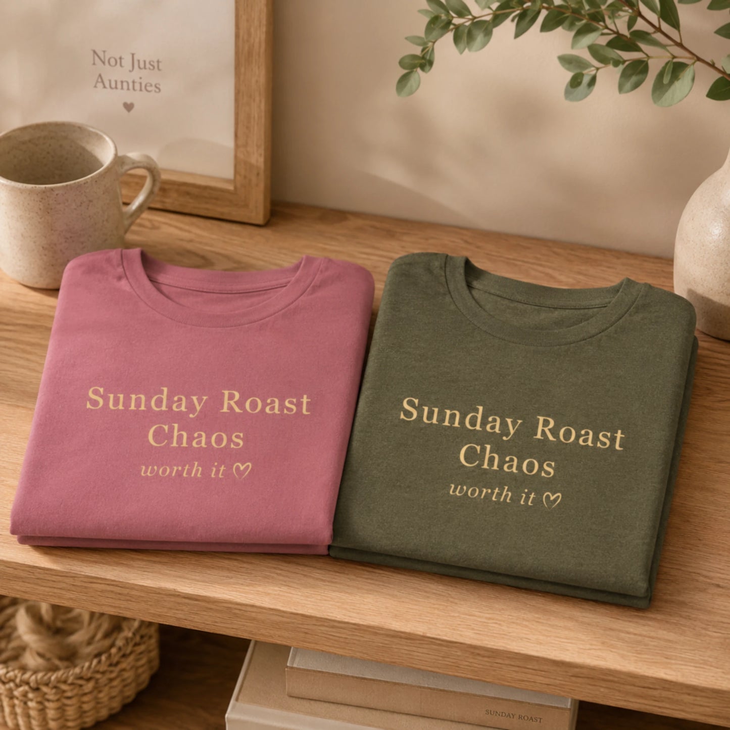 Folded mauve and olive Sunday Roast Chaos T-shirts displayed on a wooden shelf in a warm, cosy home setting.