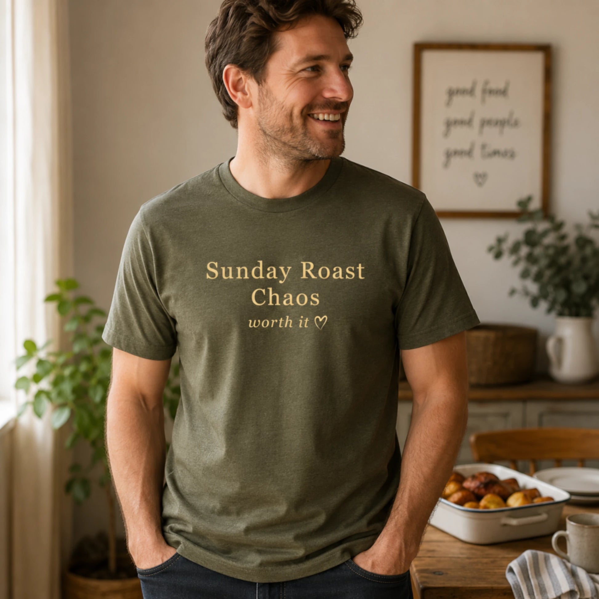 Man wearing an olive Sunday Roast Chaos T-shirt with soft cream text, standing in a relaxed, warm indoor kitchen setting.