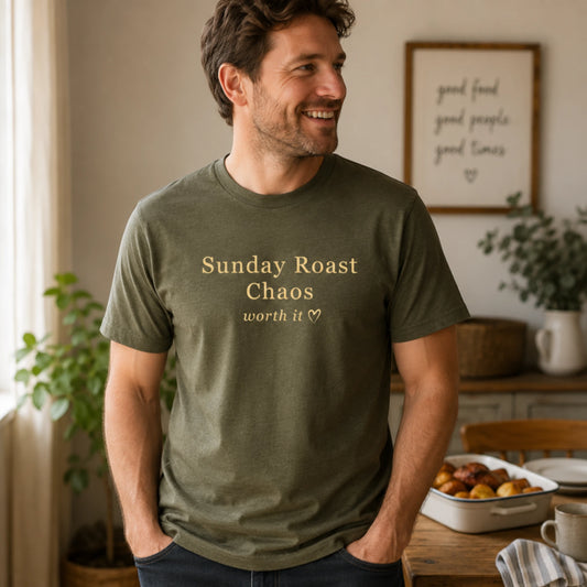 Man wearing an olive Sunday Roast Chaos T-shirt with soft cream text, standing in a relaxed, warm indoor kitchen setting.