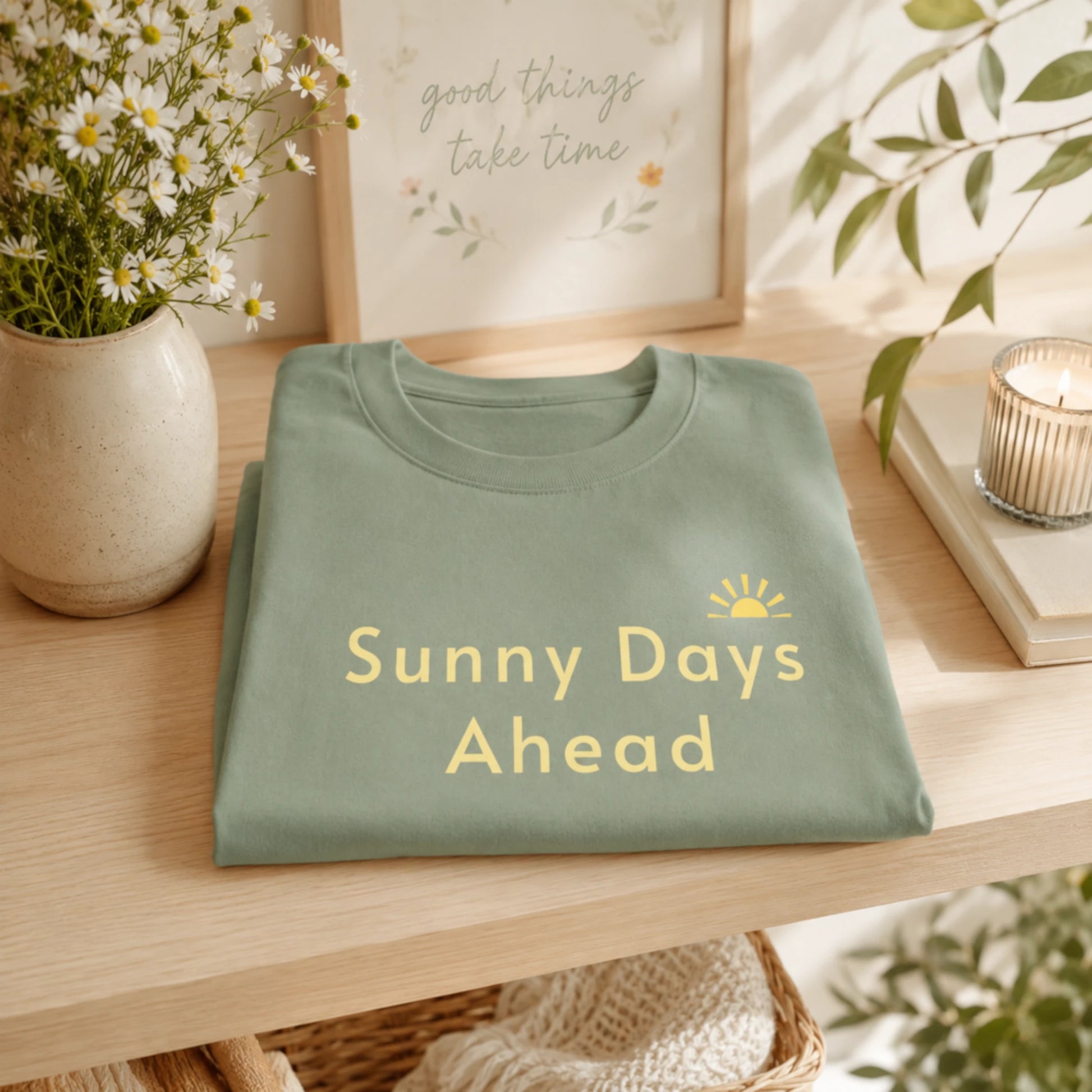 Folded Sage Sunny Days Ahead T-shirt displayed on a light wooden shelf with soft spring-inspired styling.