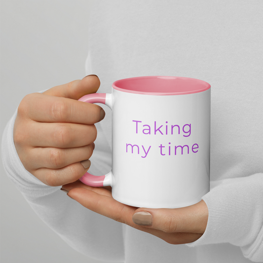 Ceramic coffee mug with pink handle and the words “taking my time”, held in hands with soft knit sleeves, capturing a relaxed and unhurried moment.