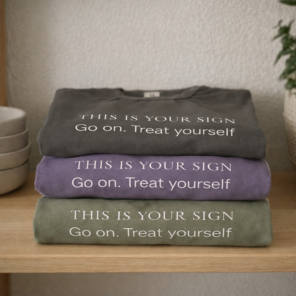 Folded stack of three graphic T-shirts in pepper, grape and moss green with “This Is Your Sign – Go on. Treat yourself” text displayed on a wooden shelf in a calm, neutral setting