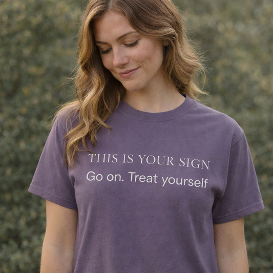 Woman wearing grape “This Is Your Sign – Go on. Treat yourself” graphic T-shirt outdoors in warm natural light with calm, relaxed expression