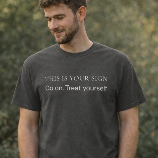 Man wearing pepper grey “This Is Your Sign – Go on. Treat yourself” graphic T-shirt outdoors in soft natural light with relaxed expression