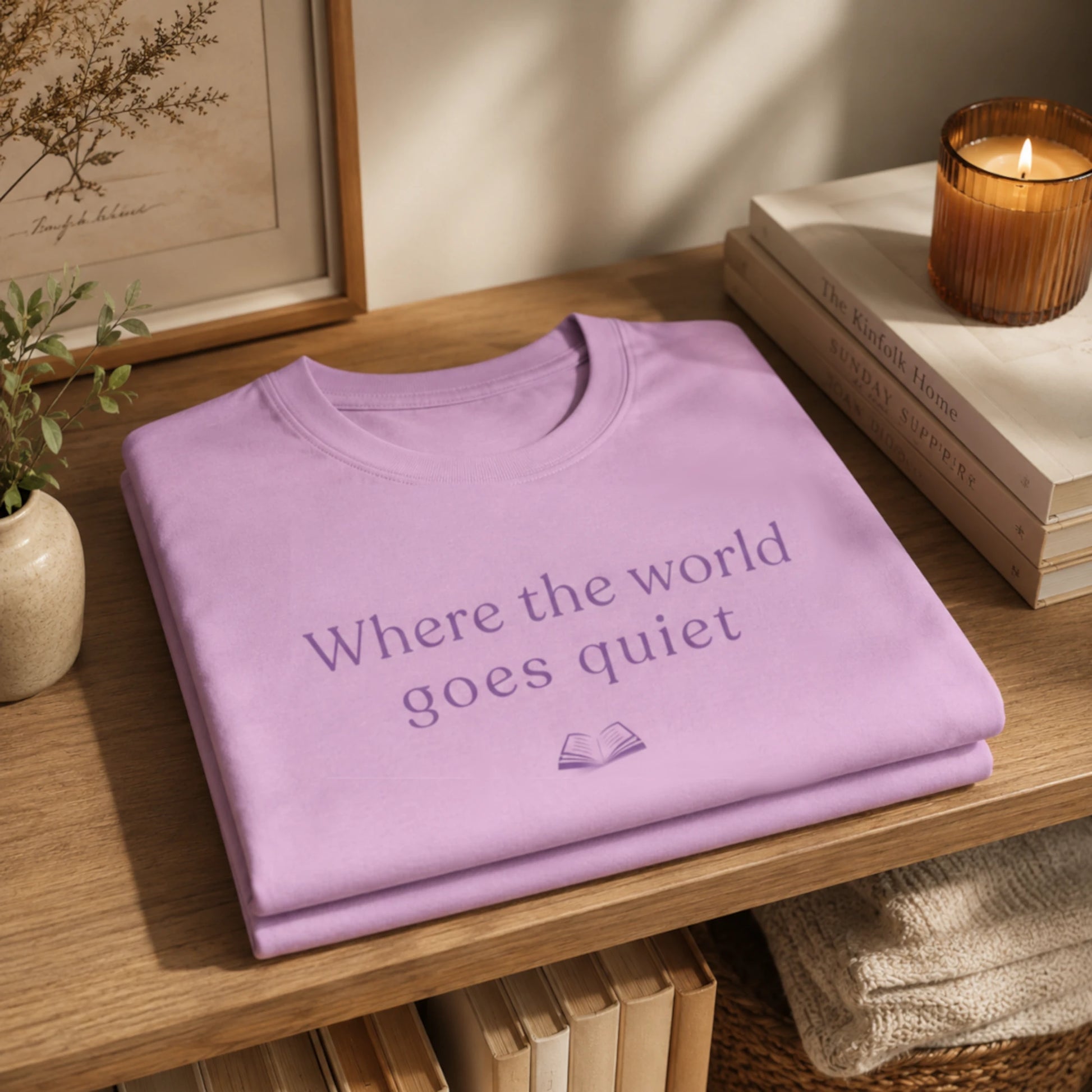 Folded Lilac “Where the World Goes Quiet” T-Shirt displayed on a wooden shelf beside stacked books and a candle in a soft, peaceful home setting.