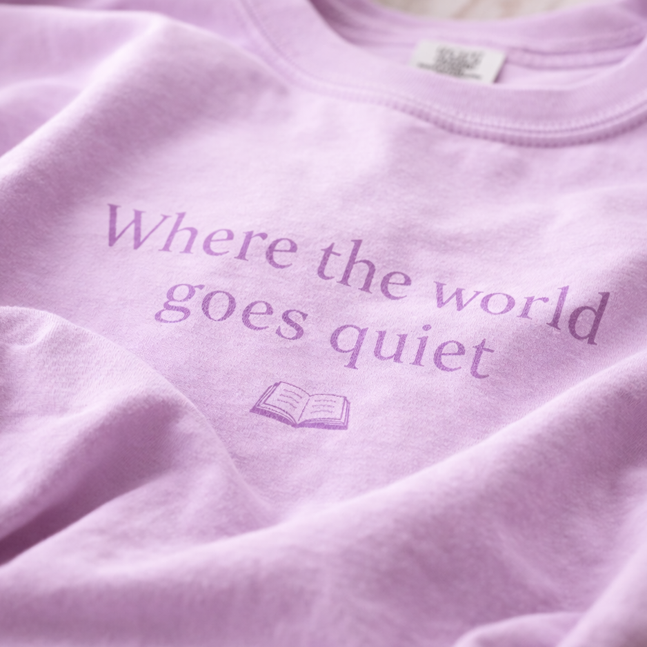 Close-up of orchid Comfort Colors T-shirt with “Where the world goes quiet” text and small open book illustration, showing soft fabric texture and relaxed folds