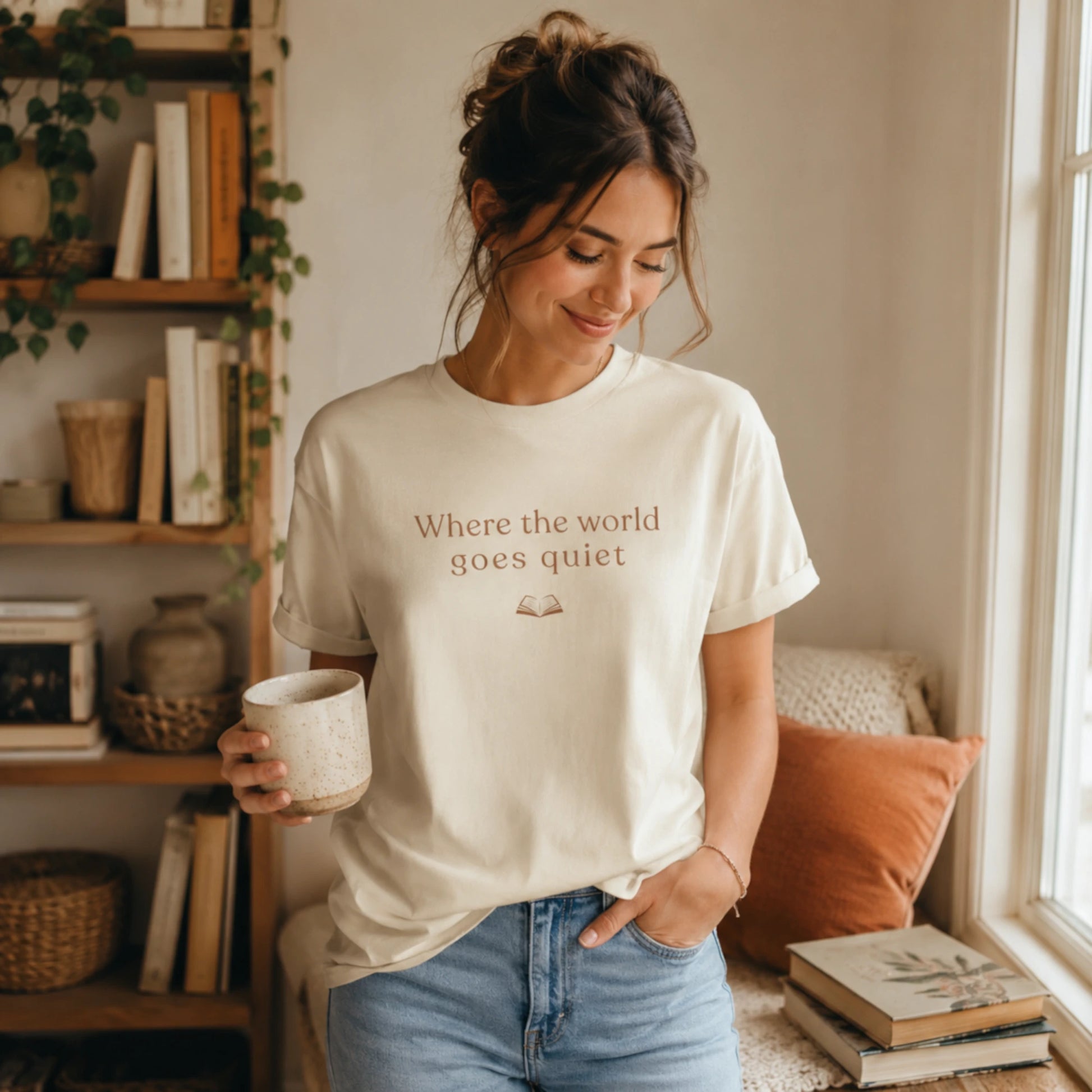 Woman wearing natural tee:
Woman wearing the Natural “Where the World Goes Quiet” T-Shirt, standing in a warm, cosy reading nook with a mug in hand and books nearby.