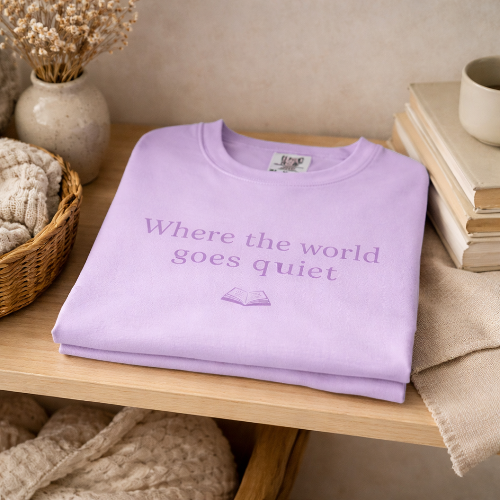 Folded orchid Comfort Colors T-shirt with “Where the world goes quiet” text displayed on wooden shelf with books and neutral decor