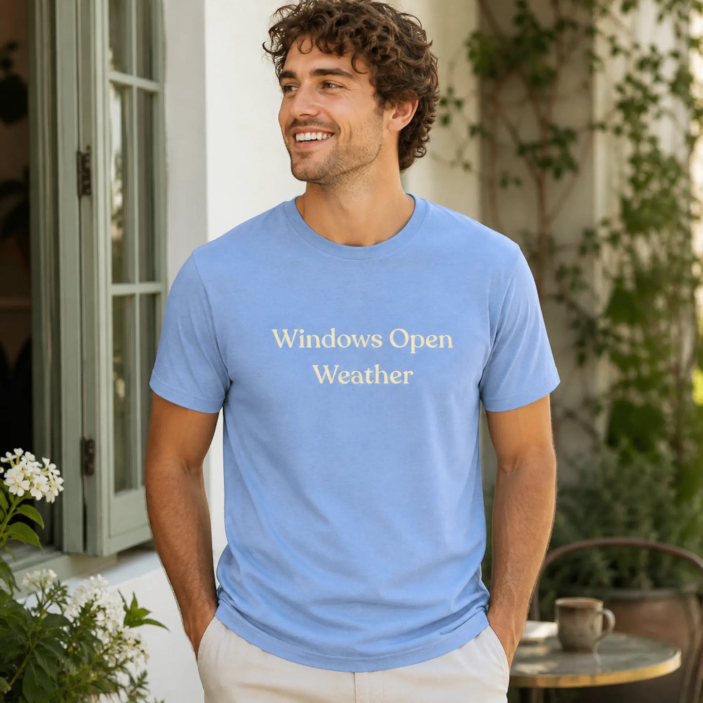 Male model wearing the Windows Open Weather T-Shirt in Heather Carolina Blue, standing outdoors in a sunny courtyard with greenery and an open window nearby.