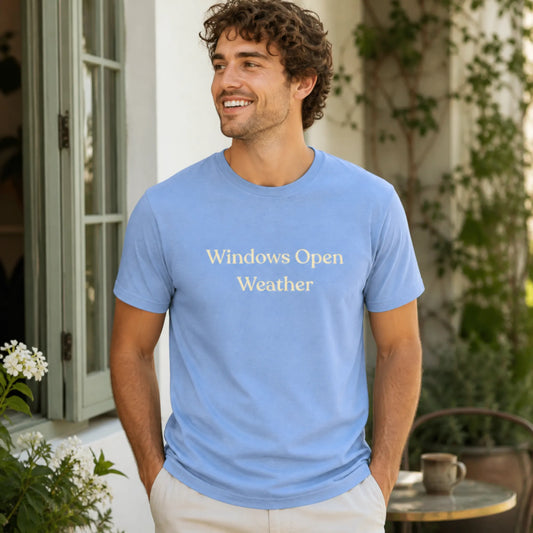 Male model wearing the Windows Open Weather T-Shirt in Heather Carolina Blue, standing outdoors in a sunny courtyard with greenery and an open window nearby.