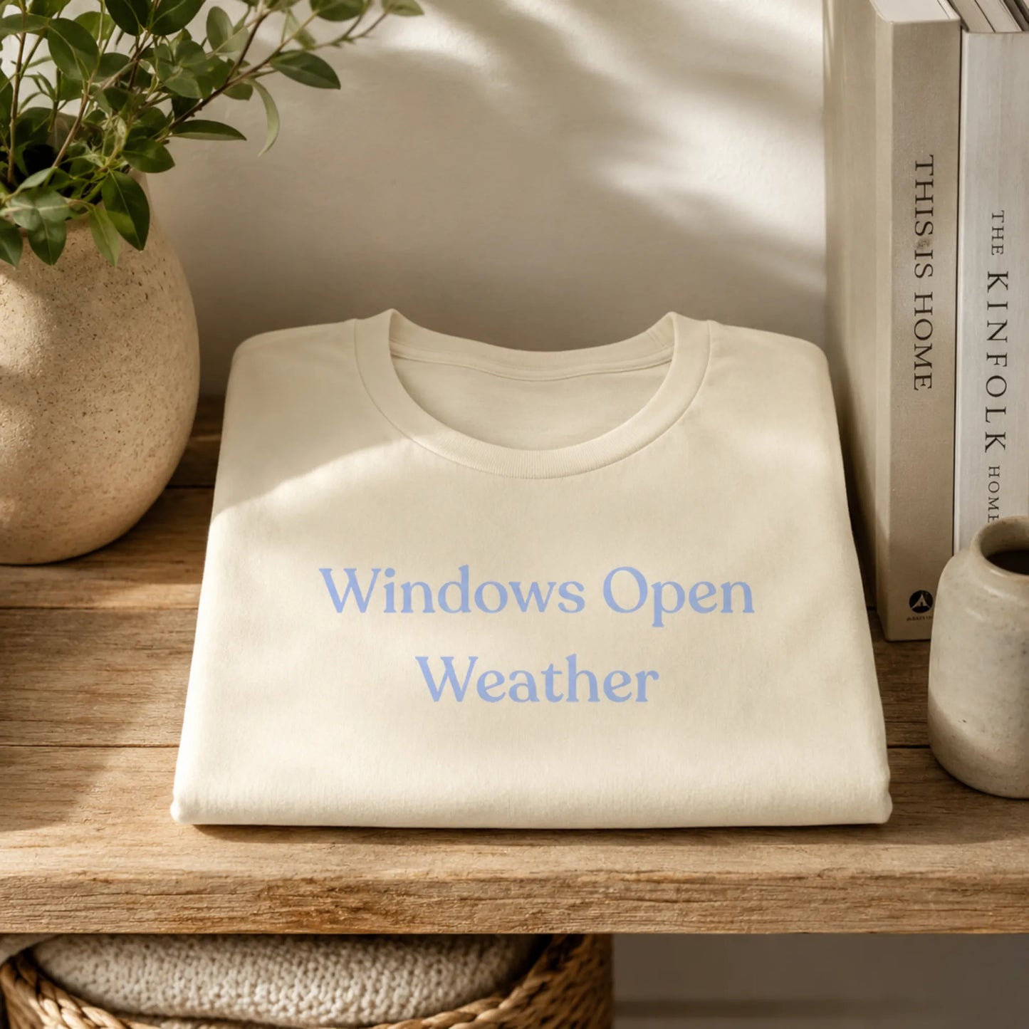 Folded Windows Open Weather T-Shirt in Natural displayed on a wooden shelf with books, a plant and soft natural light.