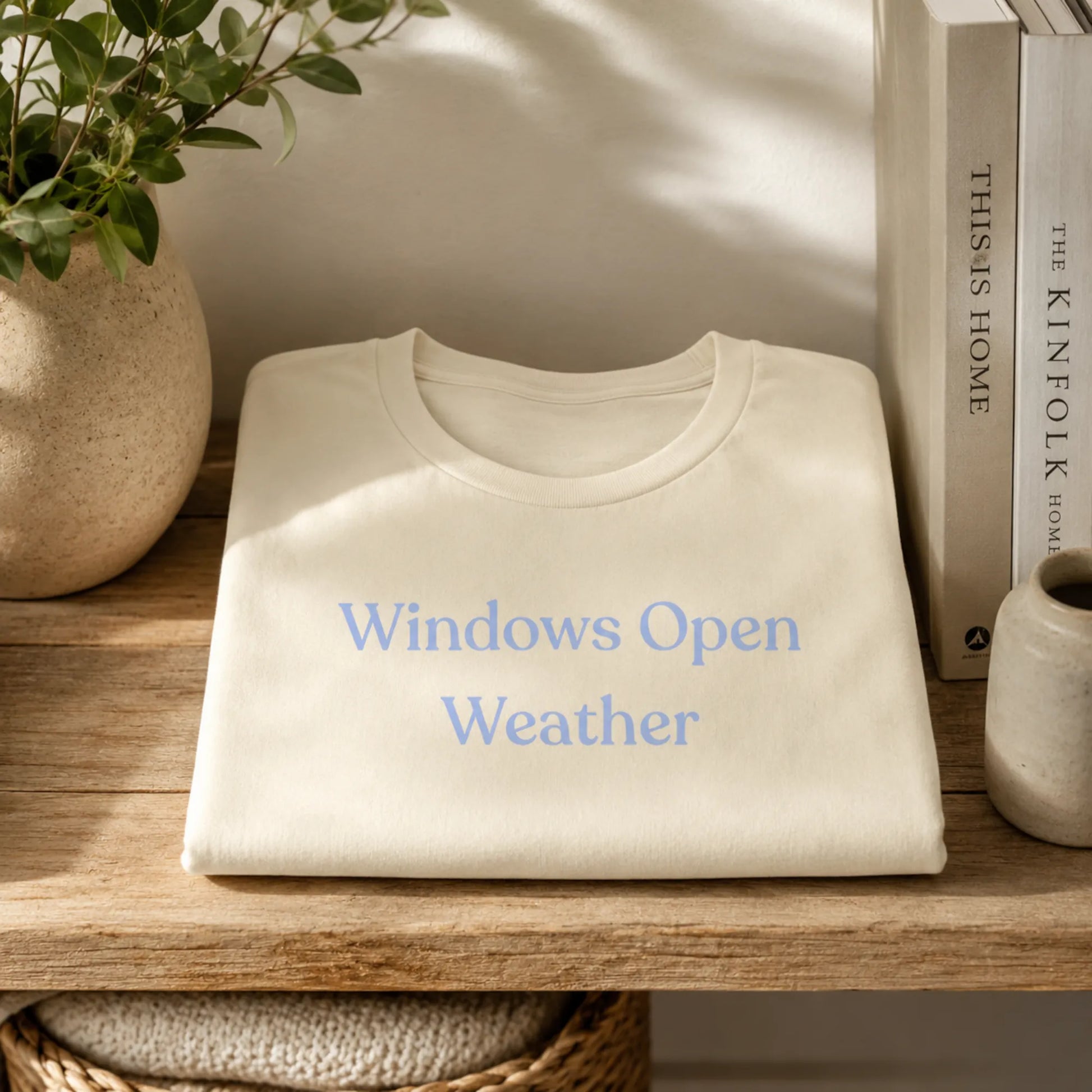 Folded Windows Open Weather T-Shirt in Natural displayed on a wooden shelf with books, a plant and soft natural light.