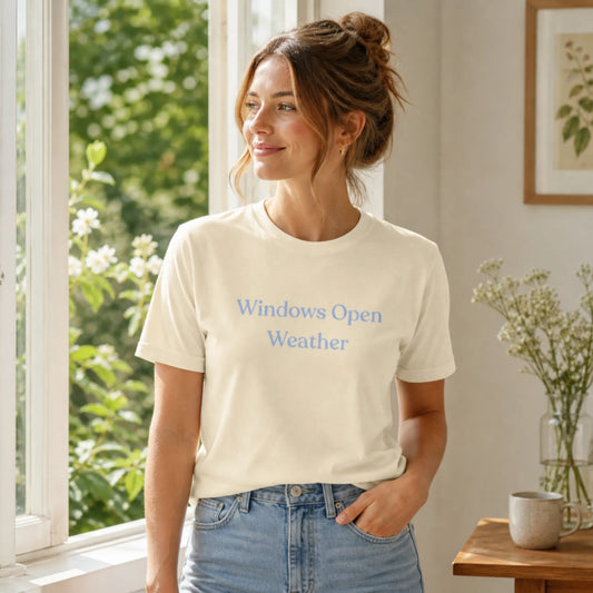 Female model wearing the Windows Open Weather T-Shirt in Natural, standing indoors beside a sunlit open window in a calm, airy room.