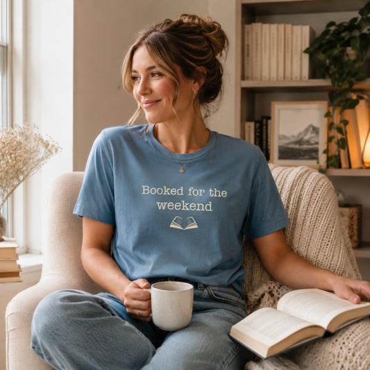 Female model wearing the steel blue “Booked for the Weekend” T-Shirt, sitting comfortably indoors with a mug in hand in a calm, cosy reading corner.