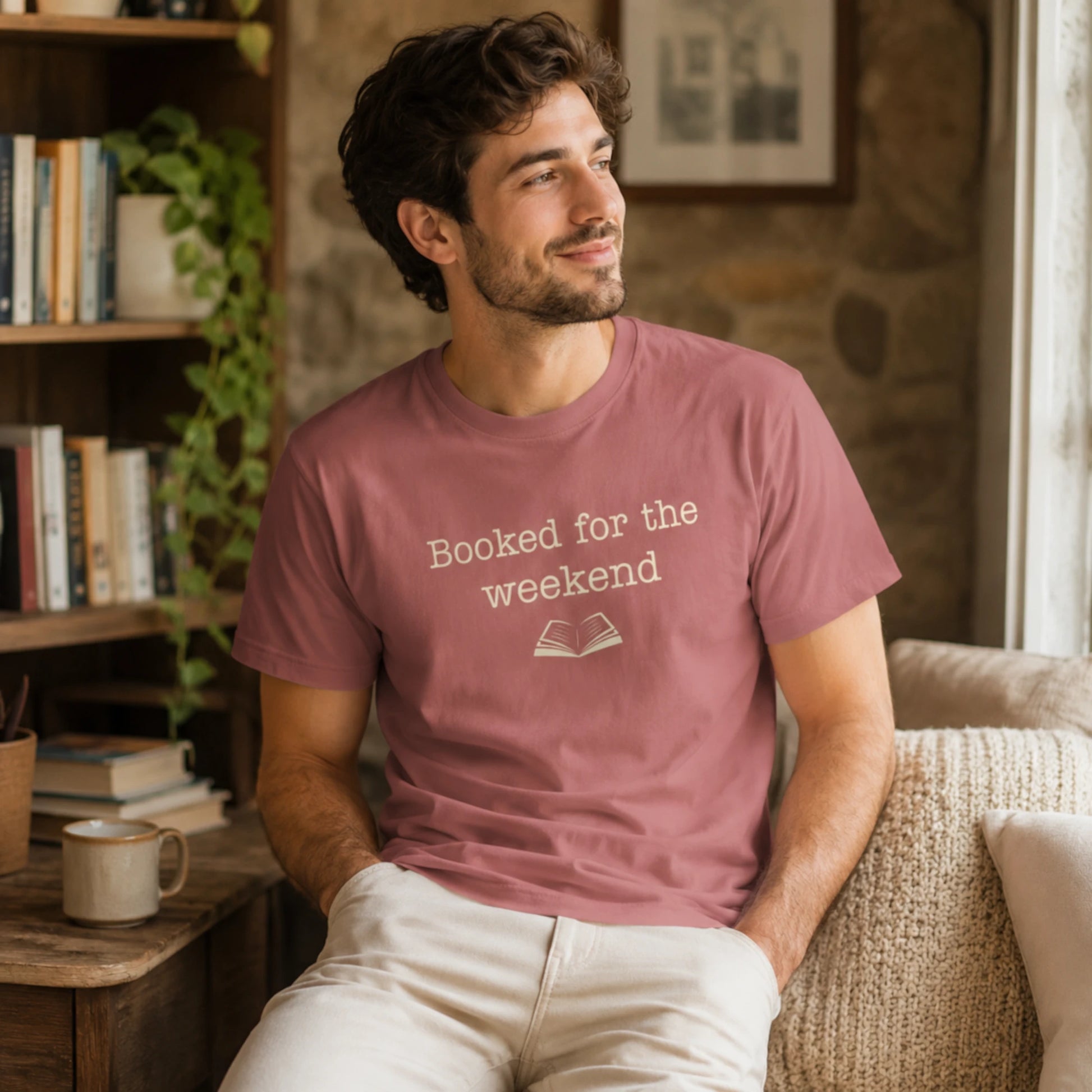 Male model wearing the mauve “Booked for the Weekend” T-Shirt, seated in a cosy indoor reading nook with soft neutral furnishings and warm natural light.