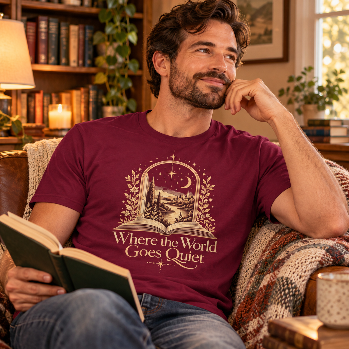 Man wearing a maroon “Where the World Goes Quiet” illustrated T-shirt while relaxing in a warm, cosy reading setting.